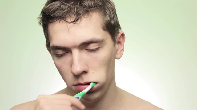 Sleepy young man is brushing his teeth with a toothbrush on an isolated light background