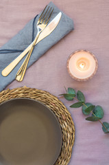 Elegant table setting on purple linen cloth dark plate rattan coaster cutlery napkin, burning candle, twig of silver dollar eucalyptus, top view