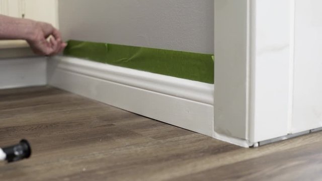 A woman as part of a DIY project carefully removes painters tape from a freshly caulked or painted baseboard