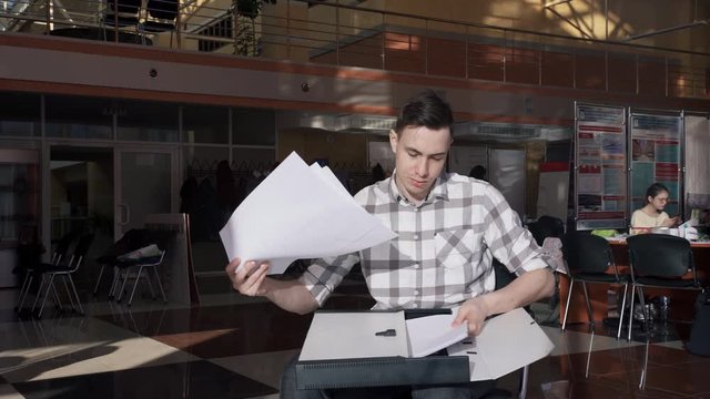 Young man sits in a public place and collects the papers in a folder. A man with short hair on her black hair is holding a pile of A4 sheets. He carefully examines the data that are on their feet. In
