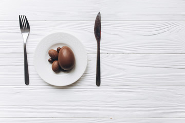 Chocolate easter eggs on plate with knife and fork on white wooden background.