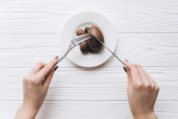 Female hands with knife and fork eating Easter chocolate eggs on white wooden background.