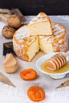 French or German soft cheese of camembert type with orange rind, honey dipper, dried fruits, knife and walnuts on wood table