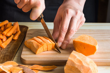 Young woman cutting with knife sweet potato into wedges, peels on wood table, sliced carrots,...