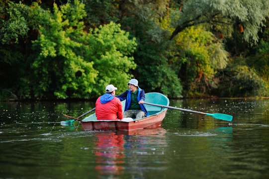 Two fishermen in a red boat with oars with fishing rods catching fish on the background of the river and nature