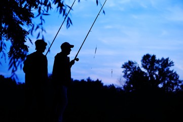 Silhouettes of two fishermen with fishing rods on nature background and blue sky. Fishing