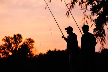 Silhouettes of two fishermen at sunset with fishing rods on the background of nature and pink sky. Fishing