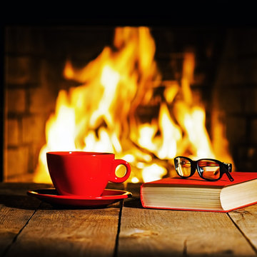 Red cup of coffee or tea, glasses and old book on wooden table near fireplace. Winter and Christmas holiday concept.