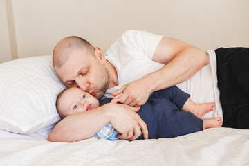 Portrait of middle age Caucasian father in white t-shirt lying in bed with newborn baby son holding his hands fingers, kissing in cheek, parenting childhood bonding concept, lifestyle