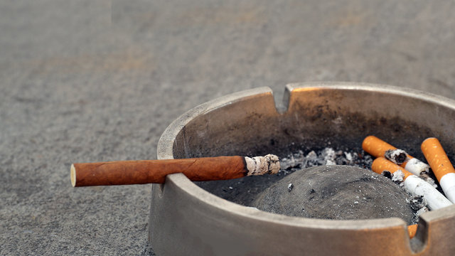 Ashtray with cigarillo and smoked cigarette butts on gray background