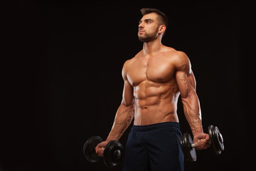 Fototapeta na wymiar Handsome athletic man in gym is pumping up muscles with dumbbells in a gym. Fitness muscular body isolated on dark background.