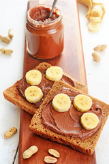Rye bread with homemade chocolate peanut butter and bananas on a wooden board over white background. Healthy breakfast close-up.