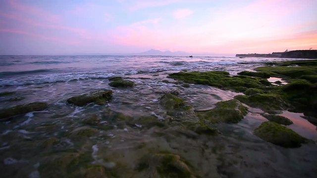 Calm ocean at sunrise, small waves wash over the rocks on the beach with mountains in the background. Bali Indonesia