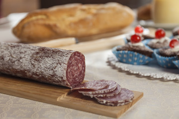 Close up of sliced sausage salame. Muffins, cheese and fresh bread in the background. Breakfast time
