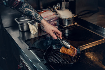 A man with tattooed arm grilling a beef in a kitchen.