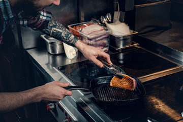 A man with tattooed arm grilling a beef in a kitchen.