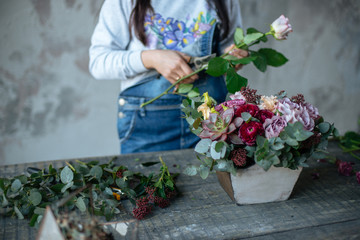 female  in gray blouse and jeans make a bouquet over gray background, putting roses in vase, flowers and vase on wood table, workplace