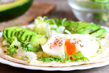 Poached egg, avocado slices, salad mix, chinese cabbage, dried herbs, sauce on a tortilla. Delicious tortilla with filling on a plate. Vegetarian breakfast. Closeup