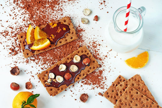 Healthy snack: homemade sandwiches with chocolate paste, hazelnut and oranges on a crispbread and jug of milk on a white table.