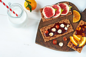 Healthy sandwiches with chocolate paste and hazelnut, soft cheese, fig, orange, on crisp rye bread and jug of milk on a white table. 