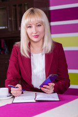 Beautiful blond woman, businesswoman, sitting in a cafe at a table, with a cup of coffee, in a burgundy suit. In the hands of a telephone, a pen and a notebook.