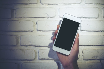 Mockup image of hand holding white mobile phone with blank black screen with white brick wall background 