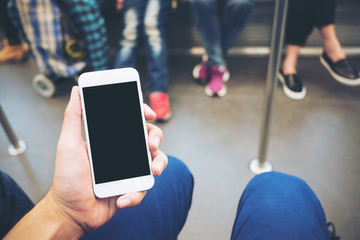 Mockup image of hand holding white mobile phone with blank black screen in subway with many people in background