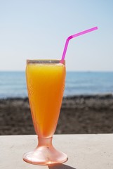 Glass of orange juice with straw with the beach and sea to the rear, Lagos, Spain.