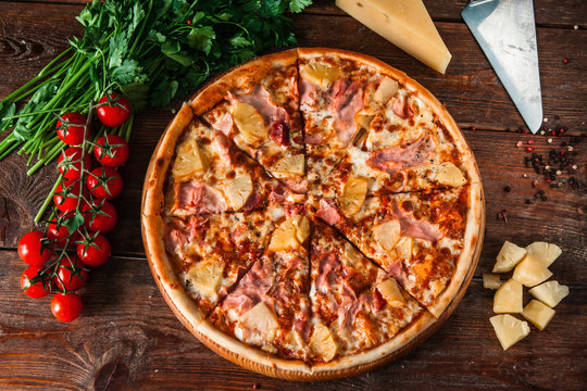 Appetizing hawaiian pizza with ham, cheese and pineapple, served on rustic wooden background with cherry, tomatoes and peppercorns, flat lay. Italian food