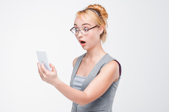 Young extremely shocked woman with mobile phone in glasses and vest on grey background. Dumbfounded woman looking surprisingly in smartphone.