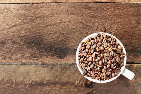 Overhead shot of a white cup filled with whole fresh coffee beans over a wood table top. Flat lay top view style.