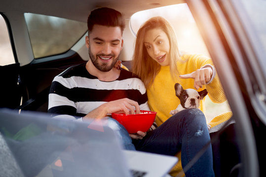 Couple Traveling By Car With Nice Doggie. Watching Movie And Eating Tasty Popcorn In Car.