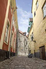 Narrow paved  stone  street in Tallinn between high houses.  motorcycle costs at  house wall - obrazy, fototapety, plakaty