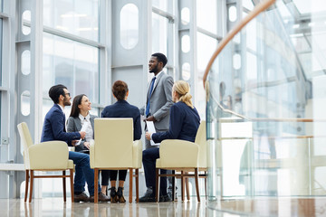 Group of  business people sitting in circle during meeting, African  man standing up giving speech  in glass hall of modern office building