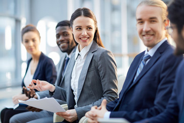 Multi-ethnic group of smiling business people sitting in row in modern glass hall, focus on young...