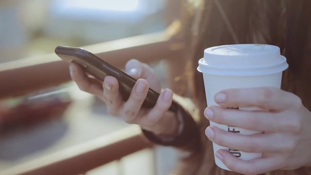 Close up of young woman using black smart phone and holding paper cup of coffee on sunset
