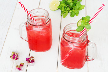Watermelon lemonade / watermelon cocktail in mason jar with red striped straw, ice cubes with rose flowers, mint and lime on a wooden white table. Selective focus.