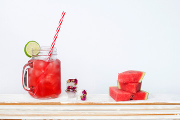 Homemade watermelon lemonade in mason jar with red striped straw, ice cubes with rose flowers and watermelon slices.
