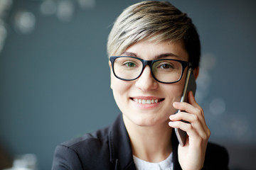 Head and shoulders portrait of smiling businesswoman in stylish eyeglasses looking at camera while calling to her colleague on smartphone