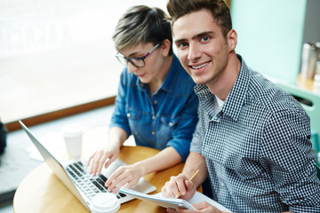 Handsome young manager looking at camera while his female colleague writing response email to their business partner