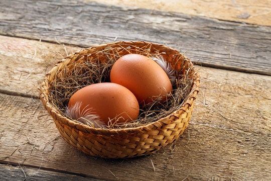 Two fresh farm eggs in a basket with hey and feather on a wooden table. Close-up shot.