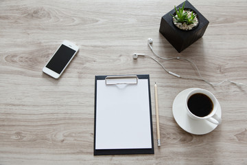 Obraz na płótnie Canvas Workplace. On the wooden table is a folder with a clip, a white sheet with space for text, pot with plant, mobile phone, headphones, a pan and white cup of coffee.