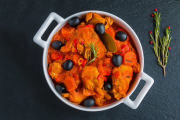 Stewed chicken,  traditional Italian dish Cacciatore in a white casserole and a rosemary on the black stone background, top view.