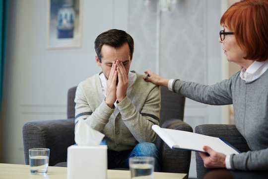Upset middle-aged patient covering face with hands while sitting on cozy armchair, his mature psychologist trying to comfort him