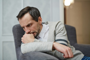 Nervous middle-aged man sitting on comfortable armchair and biting his nails while trying to find out problem solution