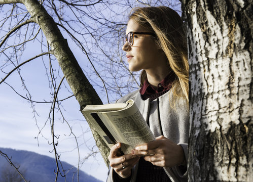 Thoughtful woman holding book while standing besides tree