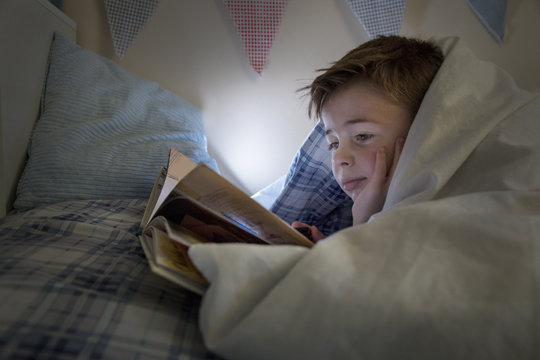 Boy lying in bed wrapped in duvet reading book