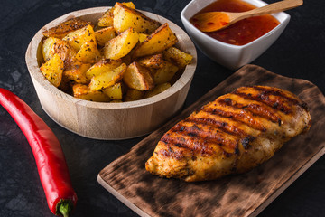 Grilled chicken breast on wooden board and grilled potato served with vegetables on black background