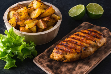 Grilled chicken breast on wooden board and grilled potato served with vegetables on black background