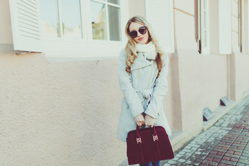 Obraz premium Happy and cheerful, smiling Blond in sunglasses young model or student in light coat on the natural street background outside.